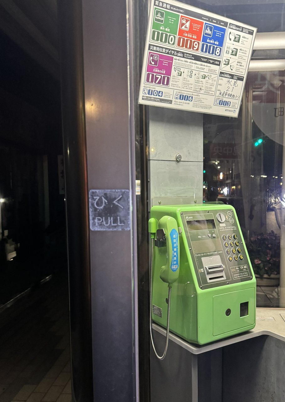 Un téléphone public vert dans une cabine téléphonique japonaise la nuit, avec au-dessus le panneau des numéros d’urgence